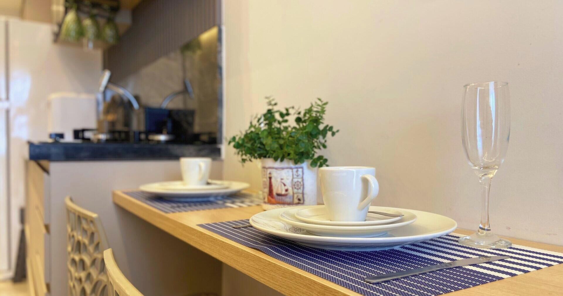 A dining area with a wooden table set with plates, cups, and a glass. A potted plant is on the wall behind the table.