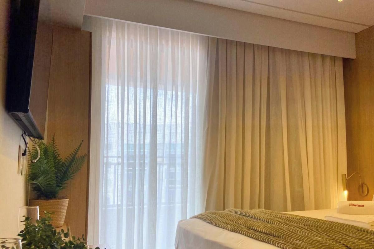 Une chambre d’hôtel avec un lit, un téléviseur fixé au mur, une grande fenêtre avec des rideaux légers et une plante en pot.