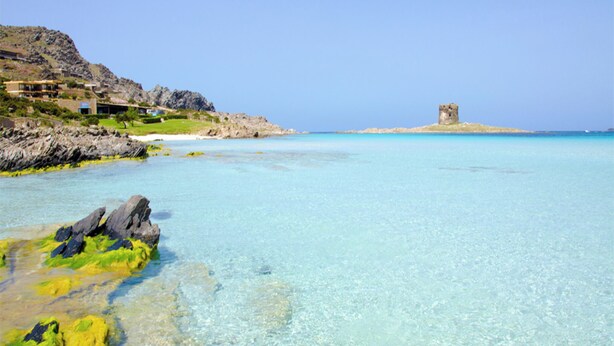 Eine Küstenlandschaft mit klarem türkisblauem Wasser, felsigen Ufern und einer in der Ferne gelegenen, schlösserähnlichen Struktur.