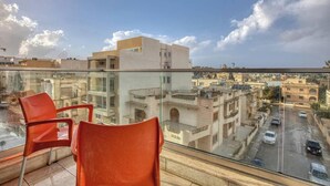 A balcony with two orange chairs, a glass railing, and a view of a cityscape with buildings and a road. - Swieqi, Central 3BR Home w/Fast WIFI & Comfort (Swieqi)