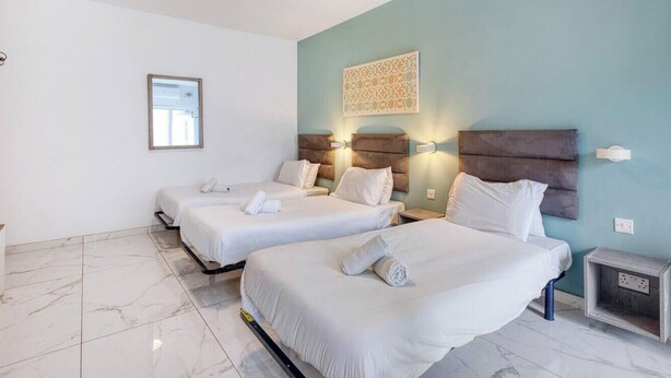 A hotel room with two beds, a small table, and a wall-mounted light fixture. - Swieqi, Central 3BR Home w/Fast WIFI & Comfort (Swieqi)