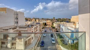 A view from a balcony overlooking a street with buildings, cars, and a clear sky. - Swieqi, Central 3BR Home w/Fast WIFI & Comfort (Swieqi)