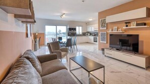 A modern living room with a sofa, a coffee table, and a TV. There is a kitchen in the background with a dining area. - Swieqi, Central 3BR Home w/Fast WIFI & Comfort (Swieqi)