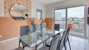 A modern dining area with a glass table and chairs, a round mirror, and a city view through large windows. - Swieqi, Central 3BR Home w/Fast WIFI & Comfort (Swieqi)