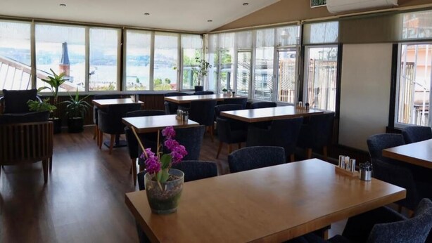 Un restaurante con varias mesas y sillas, amplias ventanas con vista a un paisaje urbano y plantas en macetas.