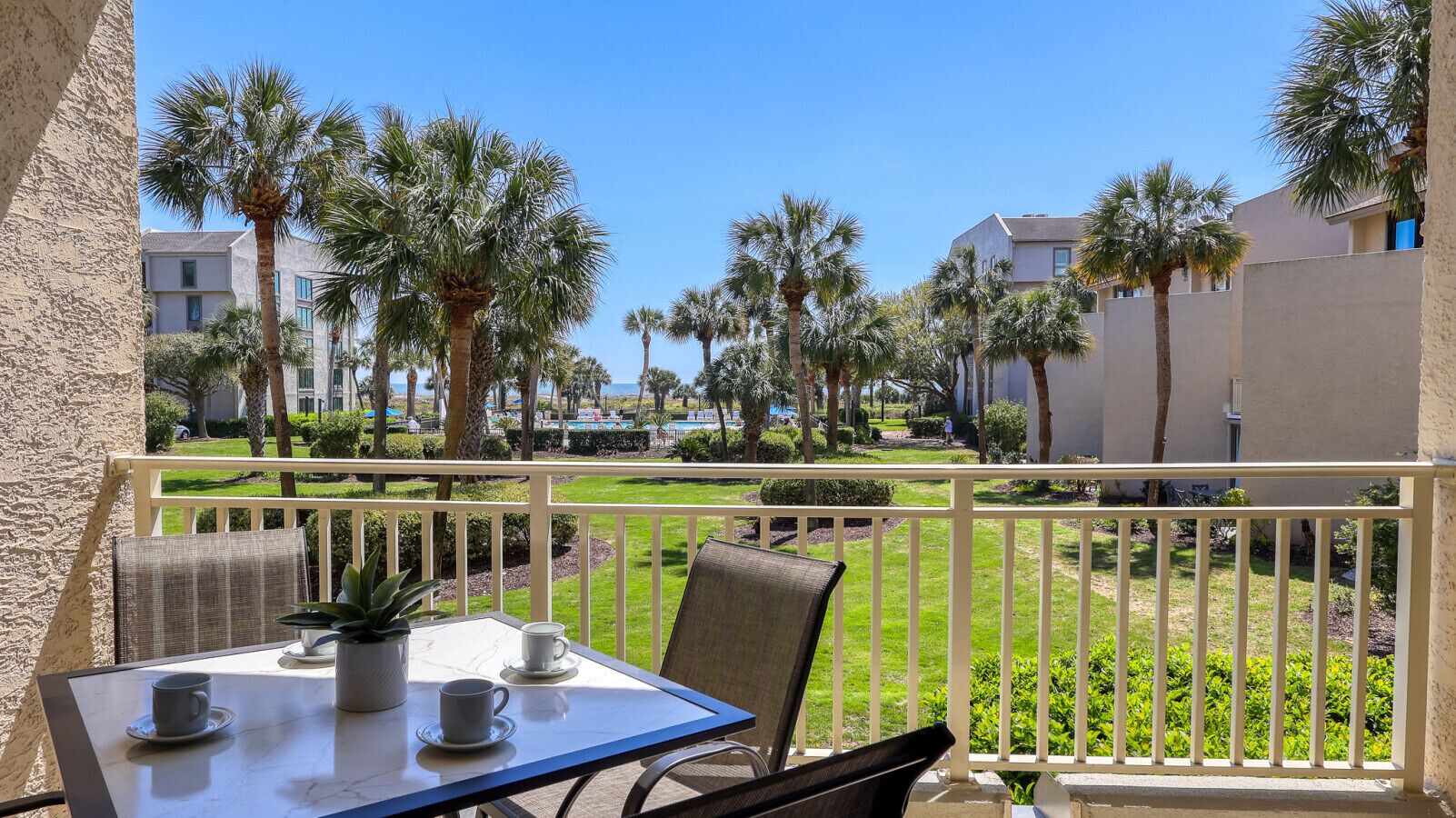 A balcony with a table and chairs, overlooking a well-maintained lawn and palm trees, with buildings in the background.