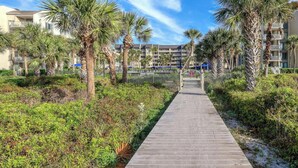 A wooden pathway leading to a building with balconies, surrounded by palm trees and greenery. - MyStay at 136 Shorewood | Ocean + Pool Views | Walk to Coligny | Sleeps 6 (Hilton Head Island)