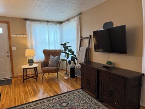 A living room with a wooden floor, a brown leather chair, a small table with a lamp, a large potted plant, a dark wooden cabinet, and a mounted TV. - Pet-Friendly Townhome • Fenced Yard • Near Avera (Sioux Falls)