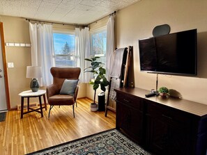Een woonkamer met een bruine leren fauteuil, een bijzettafeltje met een lamp, een grote potplant, een donker houten kast en een hangende televisie.