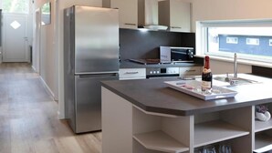 Une cuisine moderne équipée d’un réfrigérateur, d’un four à micro-ondes et d’un four en acier inoxydable, d’un petit îlot central avec une bouteille de vin, et d’une fenêtre offrant une vue agréable.
