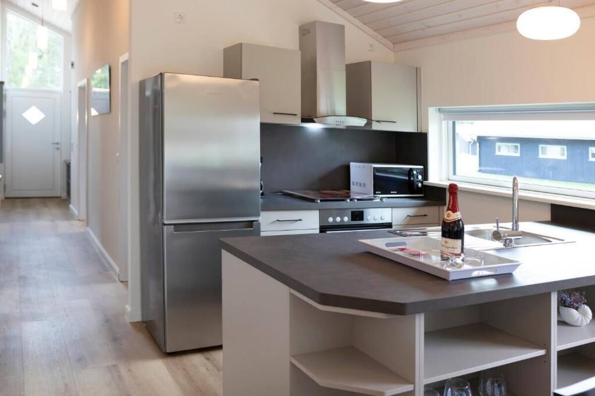 Une cuisine moderne équipée d’un réfrigérateur, d’un four à micro-ondes et d’un four en acier inoxydable, d’un petit îlot central avec une bouteille de vin, et d’une fenêtre offrant une vue agréable.