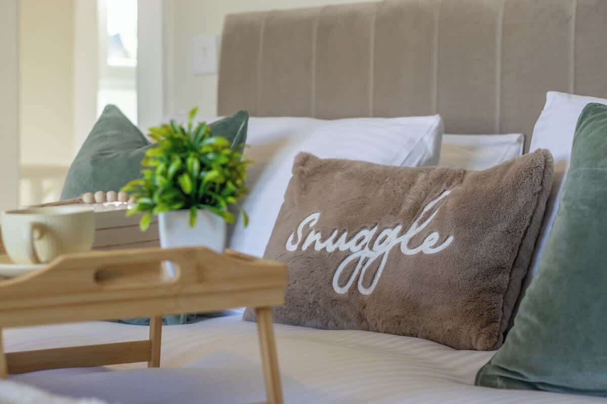 Una cama con un cojín que dice 'Snuggle', una mesita de madera y una planta en maceta.