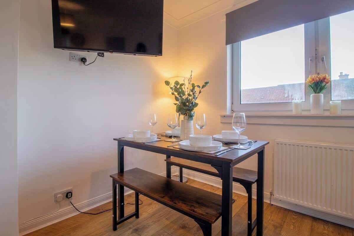 Un coin repas avec une table dressée pour deux personnes, un banc, un téléviseur fixé au mur et une fenêtre donnant sur des immeubles.