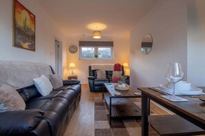 A living room with a sofa, a dining table with a wine glass, a window with a view, and a painting on the wall. - Raeburn House- 3 Bedrooms, 2 Bathrooms in Hamilton (Hamilton)