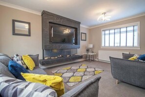 A living room with a flat-screen TV mounted on a dark wall, a fireplace, a sofa, a chair, and a window with blinds.