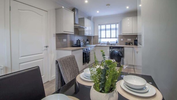 A modern kitchen with white cabinets, a dining table with plates and a potted plant, and a window providing natural light. - Catriona House - 3 Bedroom house in Motherwell (Holytown)