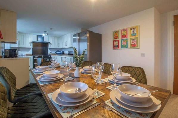 A dining area with a long table set for six, kitchen appliances in the background, and a wall with colorful framed pictures.