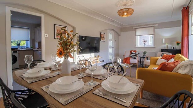 A dining area with a wooden table set for four, a vase with flowers, and a TV in the background.