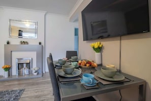 A modern dining area with a fireplace, a flat-screen TV, and a table set for two with bowls and plates. - Culzean House - 2 Bedrooms in Coatbridge (Coatbridge)