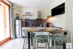 A compact kitchen with a built-in oven, a microwave, and a stainless steel refrigerator. There is a dining table with chairs and a flat-screen TV mounted on the wall. - GRABBIA 5 by Design Studio (Bellano)