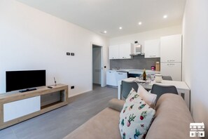 Ein modernes Wohnzimmer mit Flachbildfernseher, einem Holztischelement und einem beigefarbenen Sofa mit floralem Kissen. Im Hintergrund ist eine Küche mit weißen Schränken sowie ein Essbereich mit Tisch und Stühlen zu sehen.