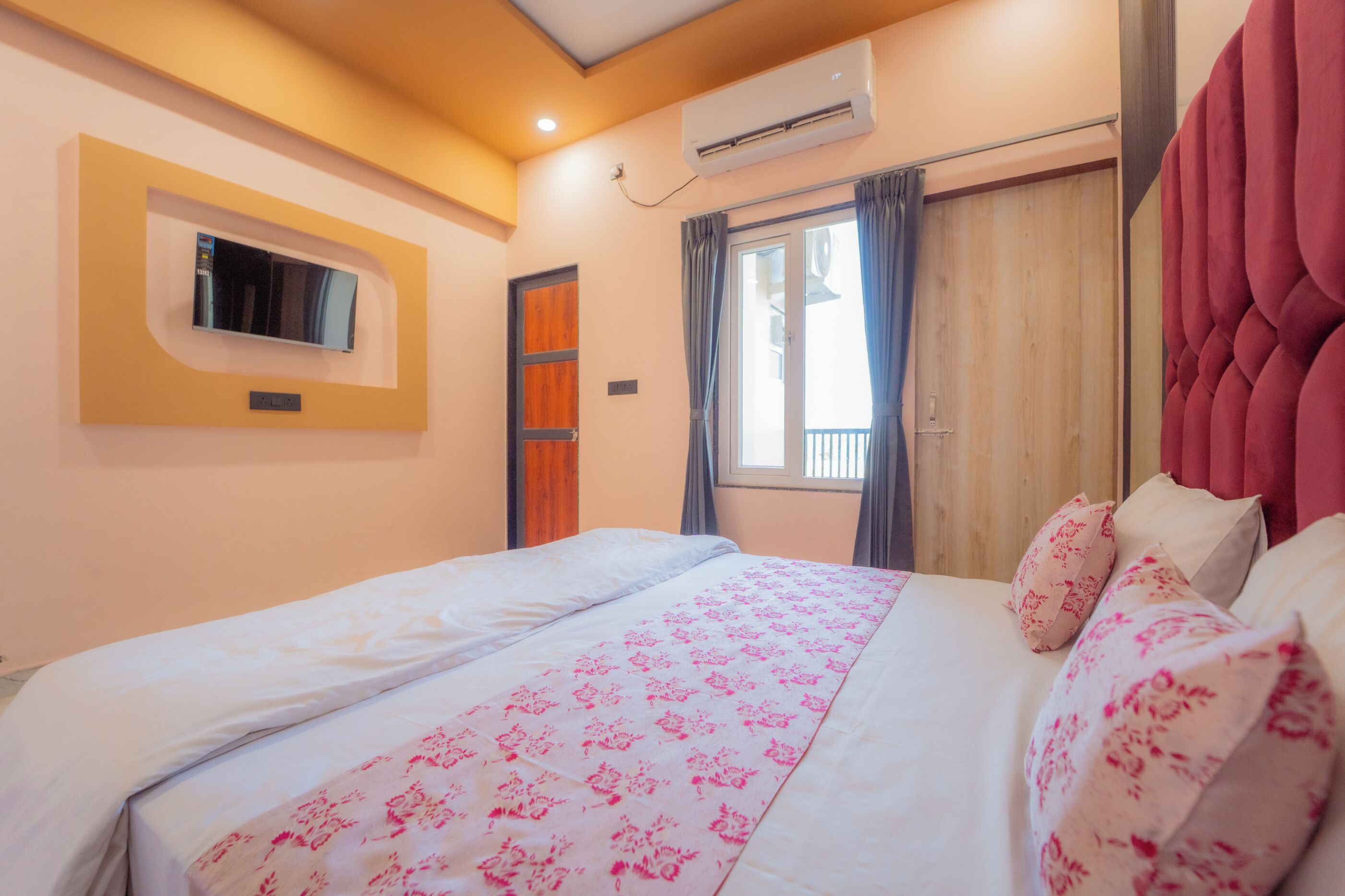 A bedroom with a bed, a TV mounted on the wall, an air conditioning unit, a window with curtains, and a wooden door.