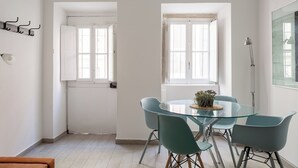 Un comedor moderno con una mesa de vidrio y sillas color esmeralda, paredes blancas y ventanas amplias.