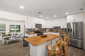 A modern kitchen with white cabinets, stainless steel appliances, a wooden island, and a dining area with wicker chairs. - Sunrise Getaway | Optional Guest House (North Fort Myers)