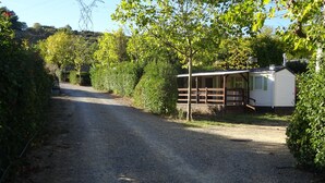 En grusvej fører hen til en lille hvid campingvogn med en indhegnet veranda, omgivet af frodig vegetation og et elmaster i baggrunden.