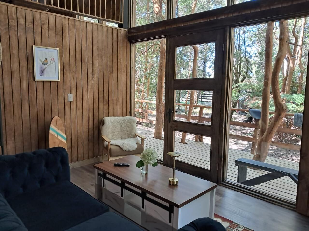Una habitación con pared de madera, una silla blanca, un sofá azul y una mesa de madera con un jarrón y un control remoto.