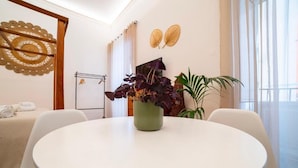 Ein modernes Zimmer mit einem runden, weißen Tisch, einer grünen Pflanze, weißen Stühlen, einem Bett mit beiger Bettdecke und einem großen Fenster mit durchscheinenden Vorhängen.