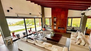 Amplia sala de estar con un sofá grande, una mesa de centro y un televisor. Amplias ventanas ofrecen vistas al exterior.