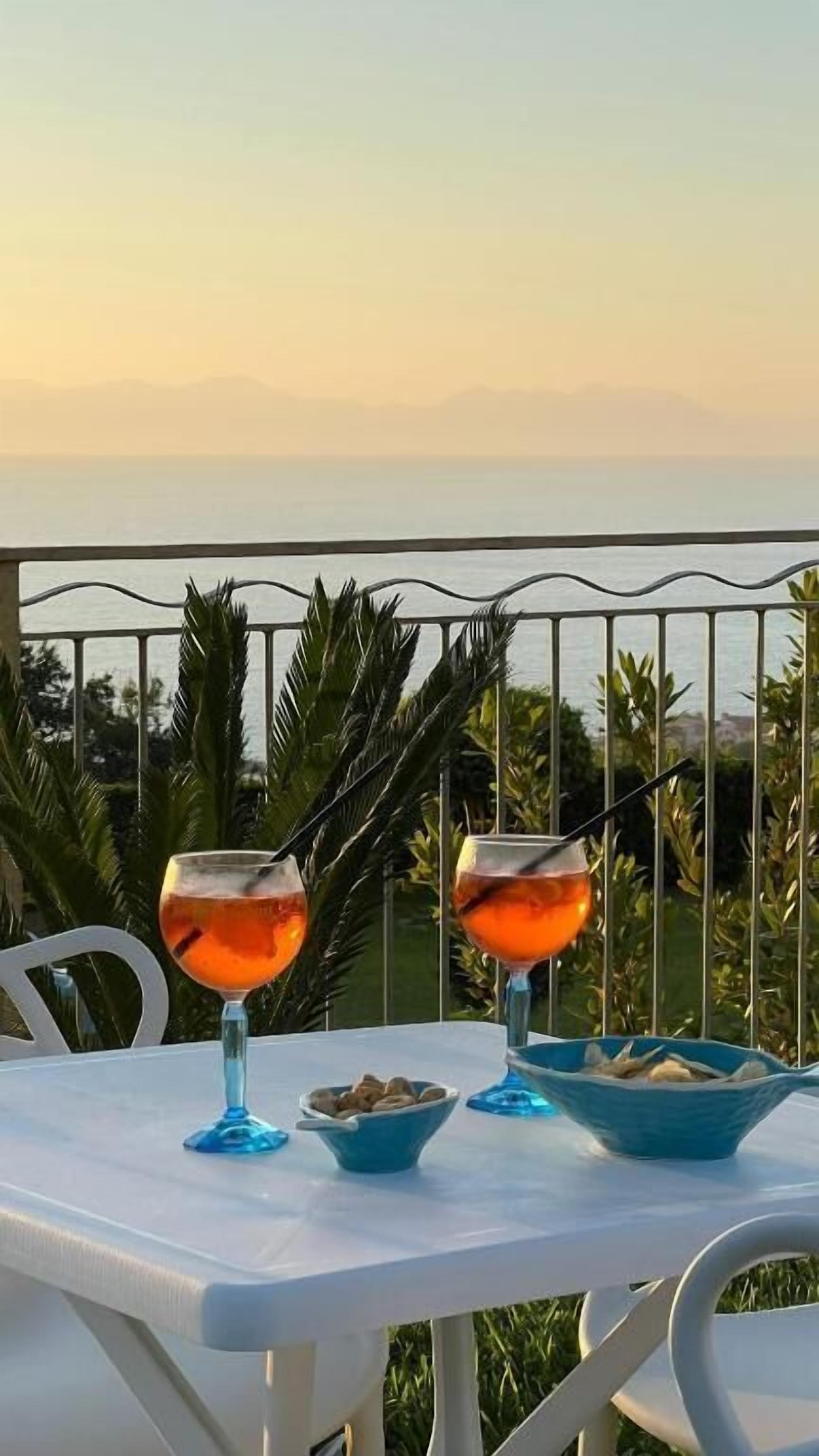 A table with two glasses of a pinkish beverage, a bowl of snacks, and a bowl of bread on a balcony overlooking a body of water.