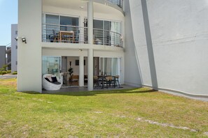 Un edificio moderno de dos pisos con un balcón, ventanas amplias y una zona de estar con una mesa y sillas.