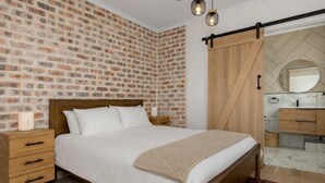 A bedroom with a brick-patterned wall, a wooden bed frame, a bedside table with a lamp, and a bathroom with a wooden door and a mirror. - Serene Green Point Stay |Quiet, Walkable and Central (Cape Town)