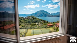 A view from a window overlooking a mountainous landscape with a lake, a tennis court, and residential buildings. - General Terms and Conditions - Vacation Rental:Welcome to our vacation rental. To ensure a pleasant and hassle-free stay, please review the following general terms and conditions.Assistance ServicesIn-person assistance: Available from 9:00 AM to 6:00 PMT (Perledo)