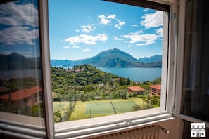 A view from a window overlooking a mountainous landscape with a lake, a tennis court, and residential buildings. - General Terms and Conditions - Vacation Rental:Welcome to our vacation rental. To ensure a pleasant and hassle-free stay, please review the following general terms and conditions.Assistance ServicesIn-person assistance: Available from 9:00 AM to 6:00 PMT (Perledo)