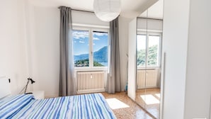 A bedroom with a bed, a window with curtains, a radiator, and a view of a mountain and lake. - General Terms and Conditions - Vacation Rental:Welcome to our vacation rental. To ensure a pleasant and hassle-free stay, please review the following general terms and conditions.Assistance ServicesIn-person assistance: Available from 9:00 AM to 6:00 PMT (Perledo)
