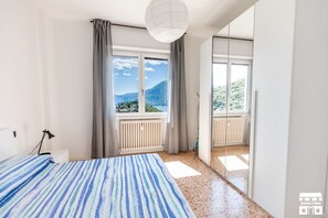 A bedroom with a bed, a window with curtains, a radiator, and a view of a mountain and lake. - General Terms and Conditions - Vacation Rental:Welcome to our vacation rental. To ensure a pleasant and hassle-free stay, please review the following general terms and conditions.Assistance ServicesIn-person assistance: Available from 9:00 AM to 6:00 PMT (Perledo)