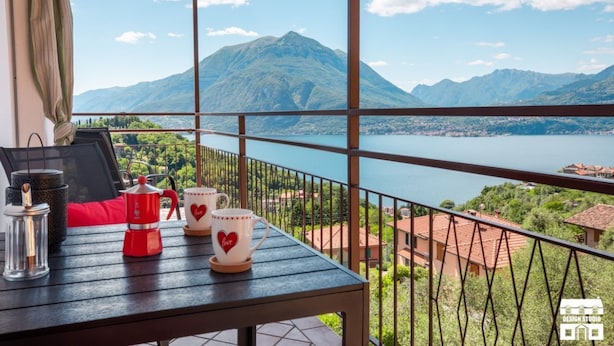 A balcony with a table, a coffee maker, and two cups with heart designs, overlooking a lake and mountains. - General Terms and Conditions - Vacation Rental:Welcome to our vacation rental. To ensure a pleasant and hassle-free stay, please review the following general terms and conditions.Assistance ServicesIn-person assistance: Available from 9:00 AM to 6:00 PMT (Perledo)