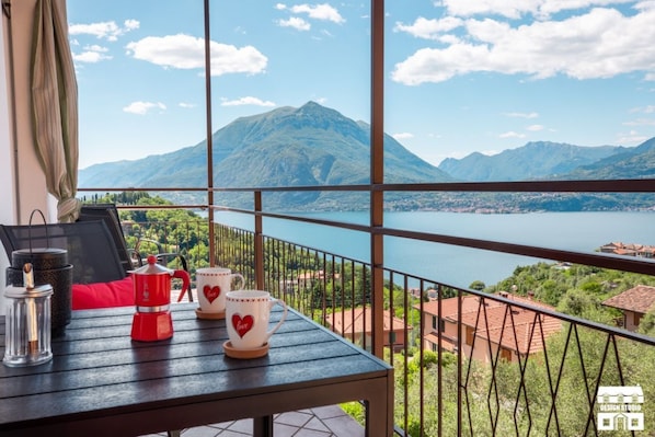 A balcony with a table, a coffee maker, and two cups with heart designs, overlooking a lake and mountains. - General Terms and Conditions - Vacation Rental:Welcome to our vacation rental. To ensure a pleasant and hassle-free stay, please review the following general terms and conditions.Assistance ServicesIn-person assistance: Available from 9:00 AM to 6:00 PMT (Perledo)