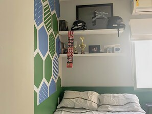 Une chambre à coucher avec un lit, une étagère remplie de livres et de trophées, et un mur orné d’un motif géométrique.
