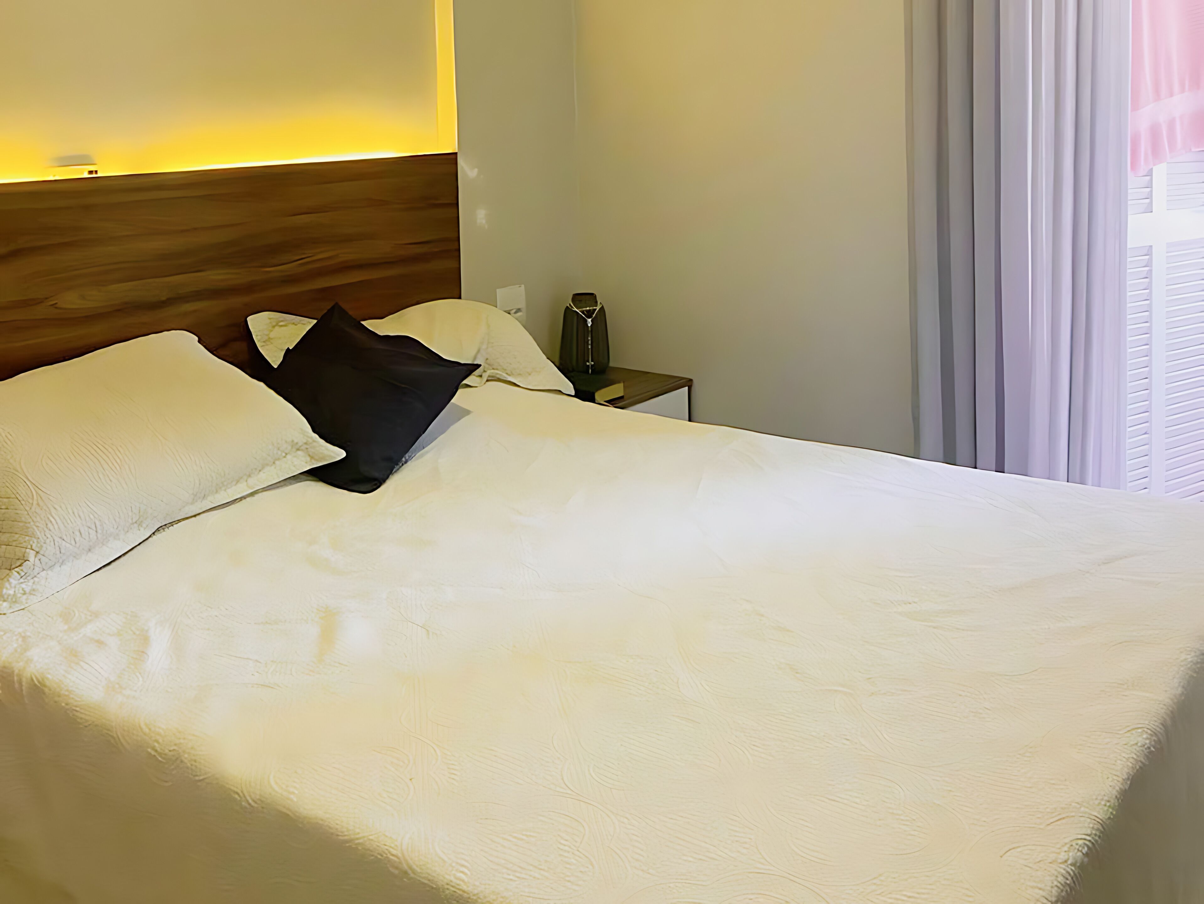 A neatly made bed with white linens and a black pillow, a wooden headboard, a bedside table with a lamp, and a window with curtains.
