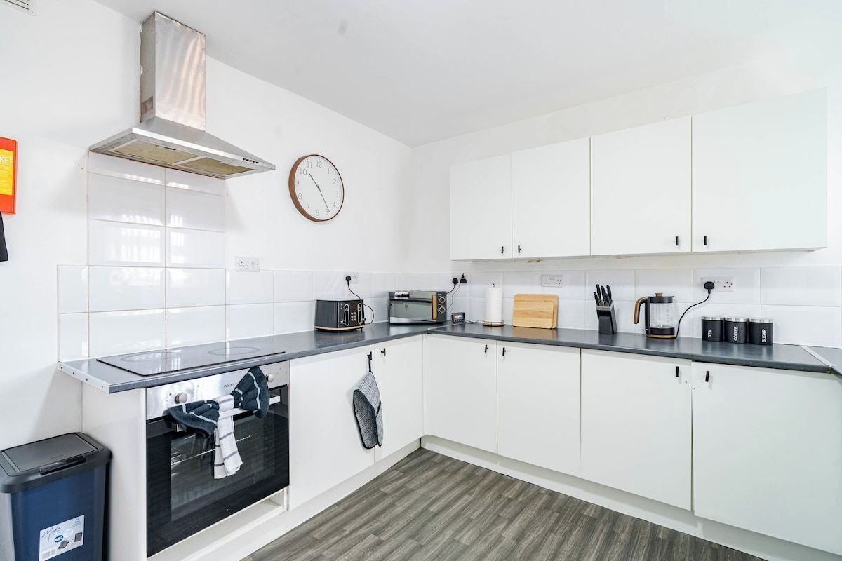 Une cuisine moderne avec des meubles blancs, un plan de travail noir, une cuisinière, un four à micro-ondes et une horloge murale.