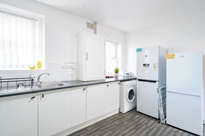 A modern kitchen with white cabinets, a built-in washing machine, a refrigerator, and a window with blinds. - Birkenhead CH43 | Spacious 6BR Home | Sleeps 12 (Birkenhead)