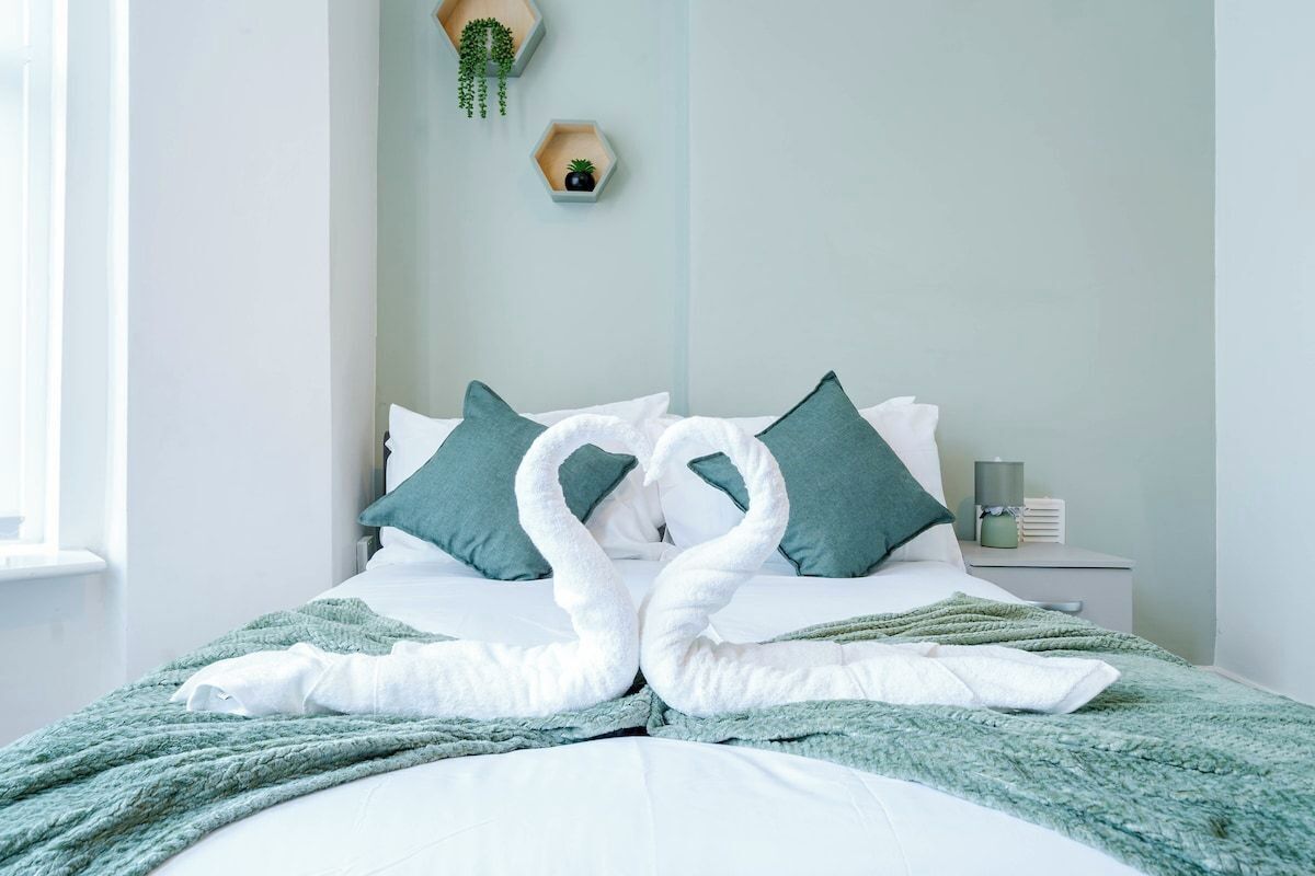 A neatly made bed with a green blanket and pillows, a bedside table with a lamp, and a wall-mounted plant.