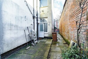 A narrow alleyway with a white wall on one side and a brick wall on the other. There is a door with a window, a ladder leaning against the white wall, and a trash bin on the ground. - Birkenhead CH43 | Spacious 6BR Home | Sleeps 12 (Birkenhead)