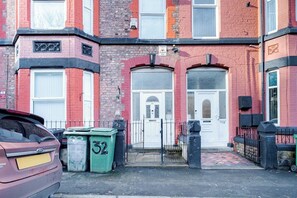 A red brick building with a white door and two windows. There are two green bins with numbers 32 and 52. - Birkenhead CH43 | Spacious 6BR Home | Sleeps 12 (Birkenhead)