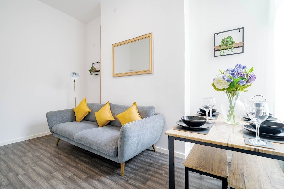 A modern living room with a grey sofa, yellow cushions, a wooden dining table set with plates and glasses, and a mirror on the wall.