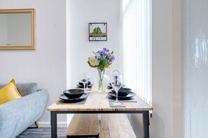 A dining area with a wooden table set for two, a vase with flowers, and a grey sofa with a yellow pillow. - Birkenhead CH43 | Spacious 6BR Home | Sleeps 12 (Birkenhead)